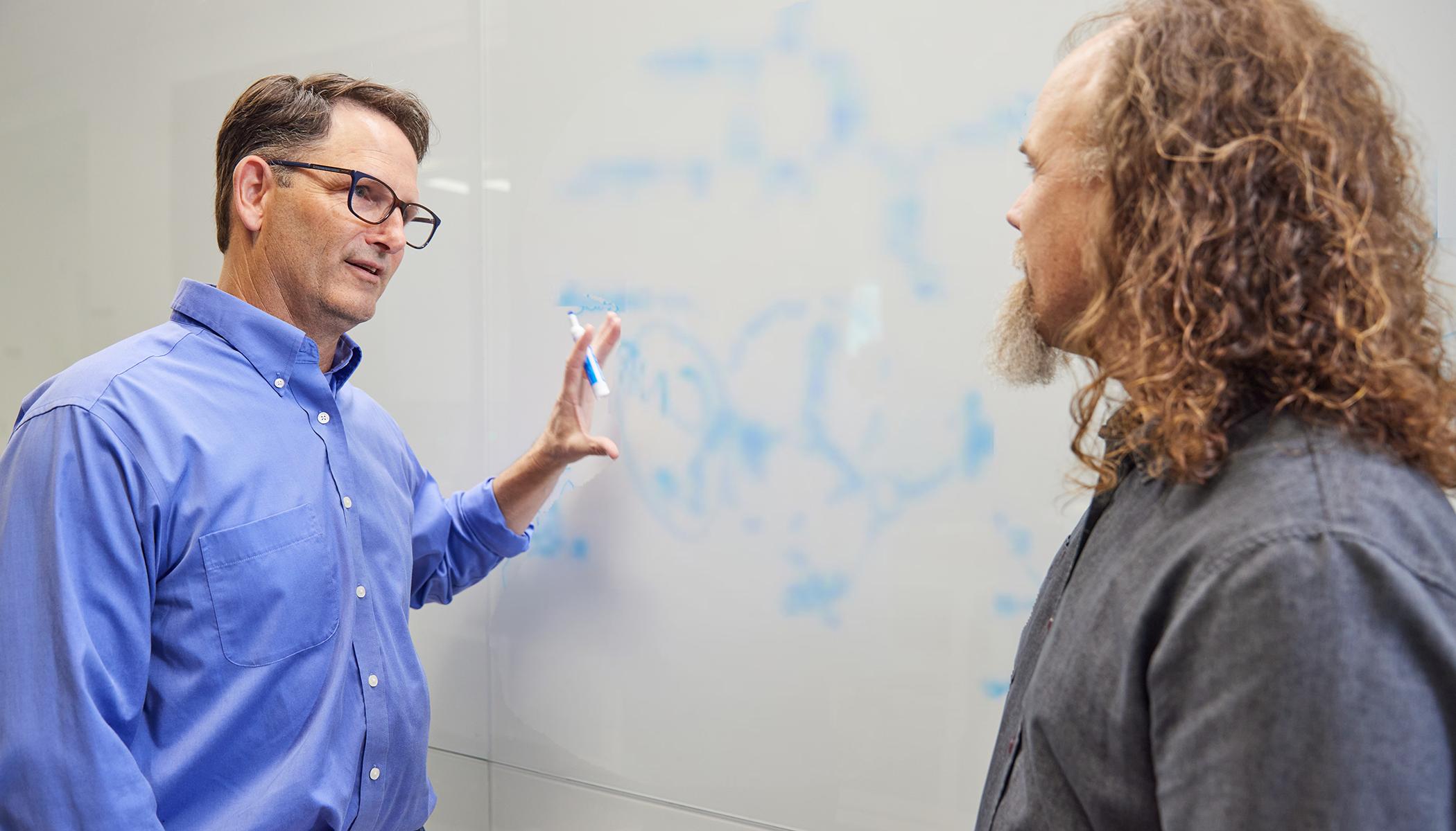 澳门正规博彩十大排行平台 scientists Brett Crawford and Dan Wendt talking by a whiteboard with a scientific formula